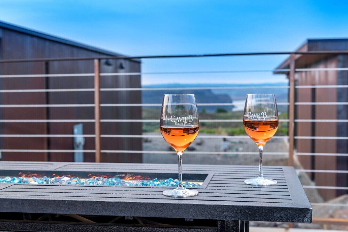 Deux verres à vin portant l’inscription « Cave B Estate Winery » sont posés sur une table, avec en arrière-plan une vue sur un bâtiment et un paysage.
