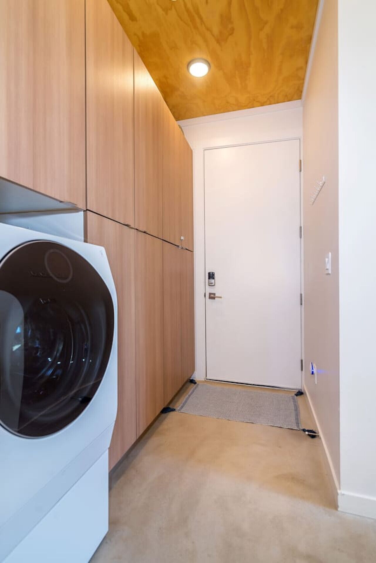 A laundry room with a front-loading washing machine, wooden cabinets, a white door, and a ceiling light.