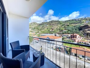A balcony with wicker furniture and a bottle of wine, overlooking a hillside with buildings. - Apartment 'Agostinho  Santa Cruz' with Sea View, Balcony and Wi-Fi (Santa Cruz)