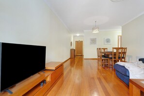 Sala de estar com televisão de tela plana, piso de madeira, área de jantar com mesa e cadeiras, e corredor que dá para outro cômodo.