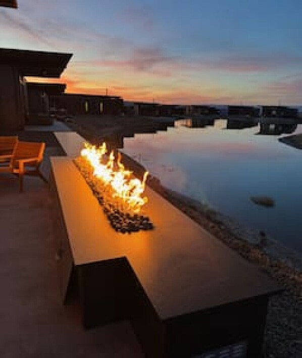 Uma longa e rectangular lareira com chamas, instalada num terraço com cadeiras e vista para um corpo de água calma e um céu ao pôr do sol.