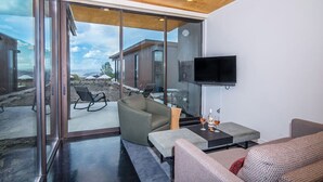A modern living room with a glass door leading to an outdoor area, a flat-screen TV mounted on the wall, and a seating area with a small table.