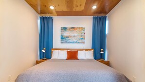 Une chambre à coucher avec une tête de lit en bois, un grand lit recouvert d’une couverture grise, deux lampes de chevet et un rideau bleu.