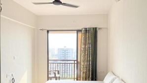 Et hotelværelse med seng, ventilator, et vindue med gardiner og udsigt til bygninger.