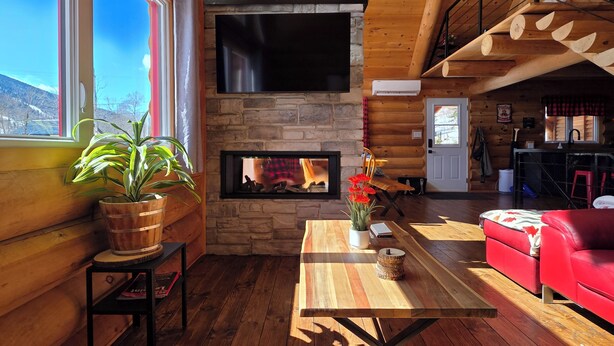 A log cabin interior with a stone fireplace, a television mounted above it, a red sofa, a wooden dining table, and a large window with a view of the mountains. - The Sugar Camp | Spa. Log cabin. Southern Massif. (Saint-Philémon)