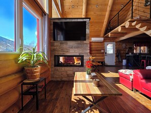 A log cabin interior with a stone fireplace, a television mounted above it, a red sofa, a wooden dining table, and a large window with a view of the mountains. - The Sugar Camp | Spa. Log cabin. Southern Massif. (Saint-Philémon)
