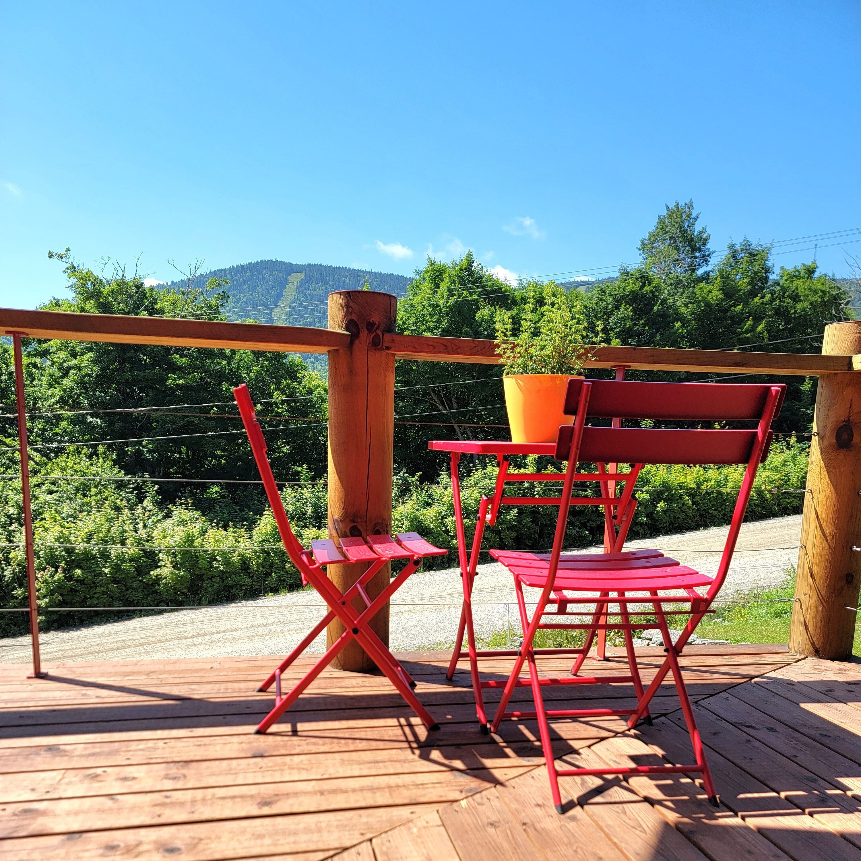 A wooden deck with red chairs and a table, overlooking a forested area with a mountain in the background.