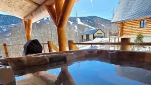 A hot tub with wooden beams overhead, a wooden cabin in the background, and a snowy mountain view. - The Sugar Camp | Spa. Log cabin. Southern Massif. (Saint-Philémon)