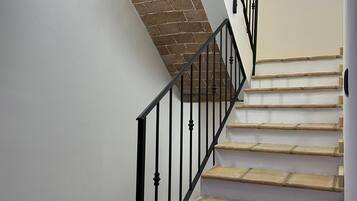 Uma escadaria com degraus de madeira e corrimãos de metal preto, que leva a um quarto com paredes brancas e uma parede de destaque em tijolo.