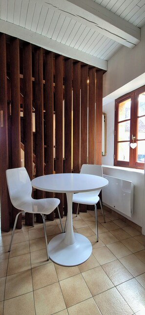 A dining area with a round table and two chairs, a wooden partition, a window with a view, and a radiator. - Cozy house in the heart of the Bastide with private courtyard and closed garage (Domme)