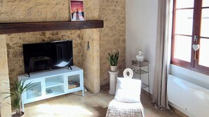 A living room with a stone wall, wooden ceiling beams, a flat-screen TV on a white stand, a sofa, a round woven rug, and a window with curtains. - Cozy house in the heart of the Bastide with private courtyard and closed garage (Domme)