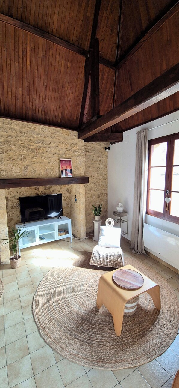 A living room with a stone wall, wooden ceiling beams, a flat-screen TV on a white stand, a sofa, a round woven rug, and a window with curtains. - Cozy house in the heart of the Bastide with private courtyard and closed garage (Domme)
