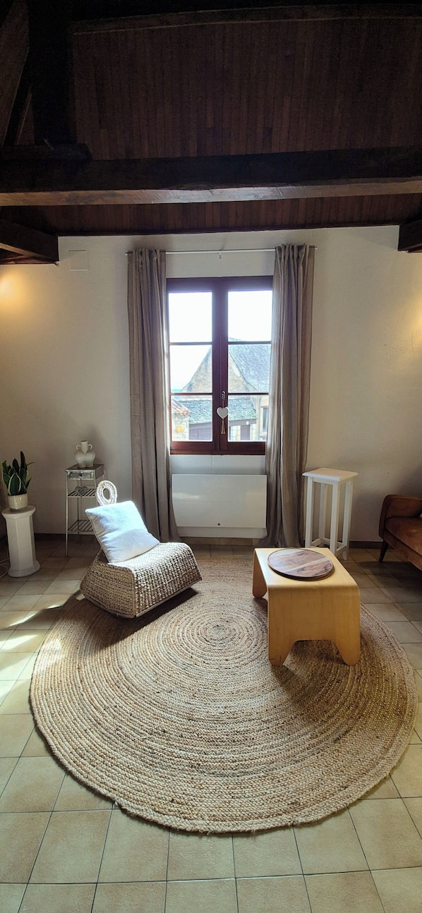 A cozy living room with a large window, a woven chair, a small wooden stool, and a potted plant. - Cozy house in the heart of the Bastide with private courtyard and closed garage (Domme)