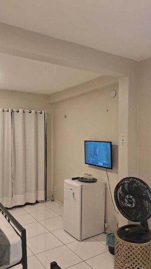A room with a bed, a small white refrigerator, a television mounted on the wall, and a standing fan. - Pousada Vasconcelos Tiangua (Tianguá)