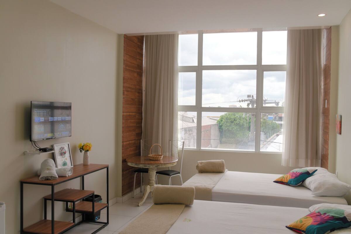 A hotel room with two beds, a TV, a desk with a chair, a window with curtains, and a view of the outside.
