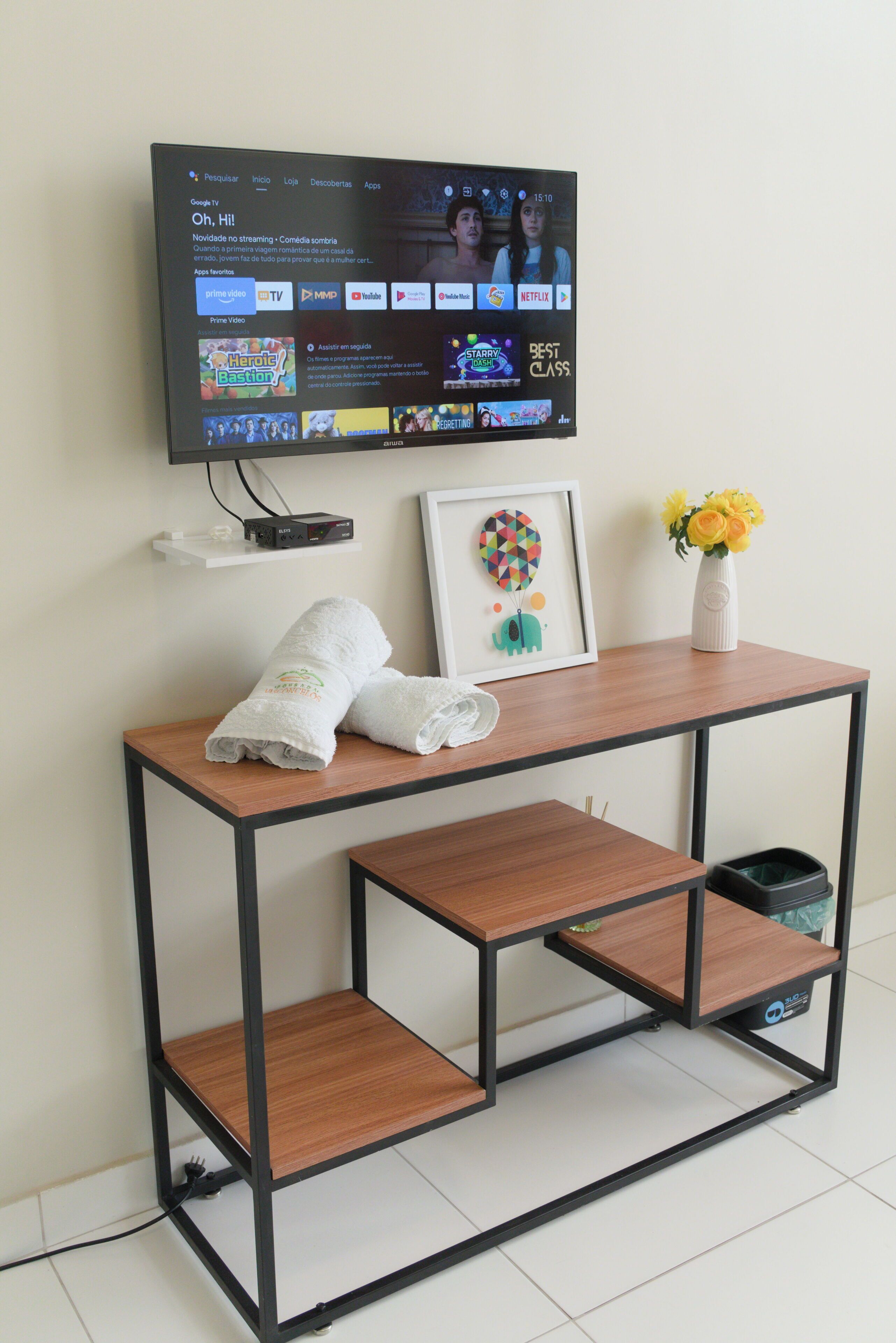 A modern room with a TV mounted on the wall, a wooden table with a white vase and a framed picture, and a small shelf with a trash bin.