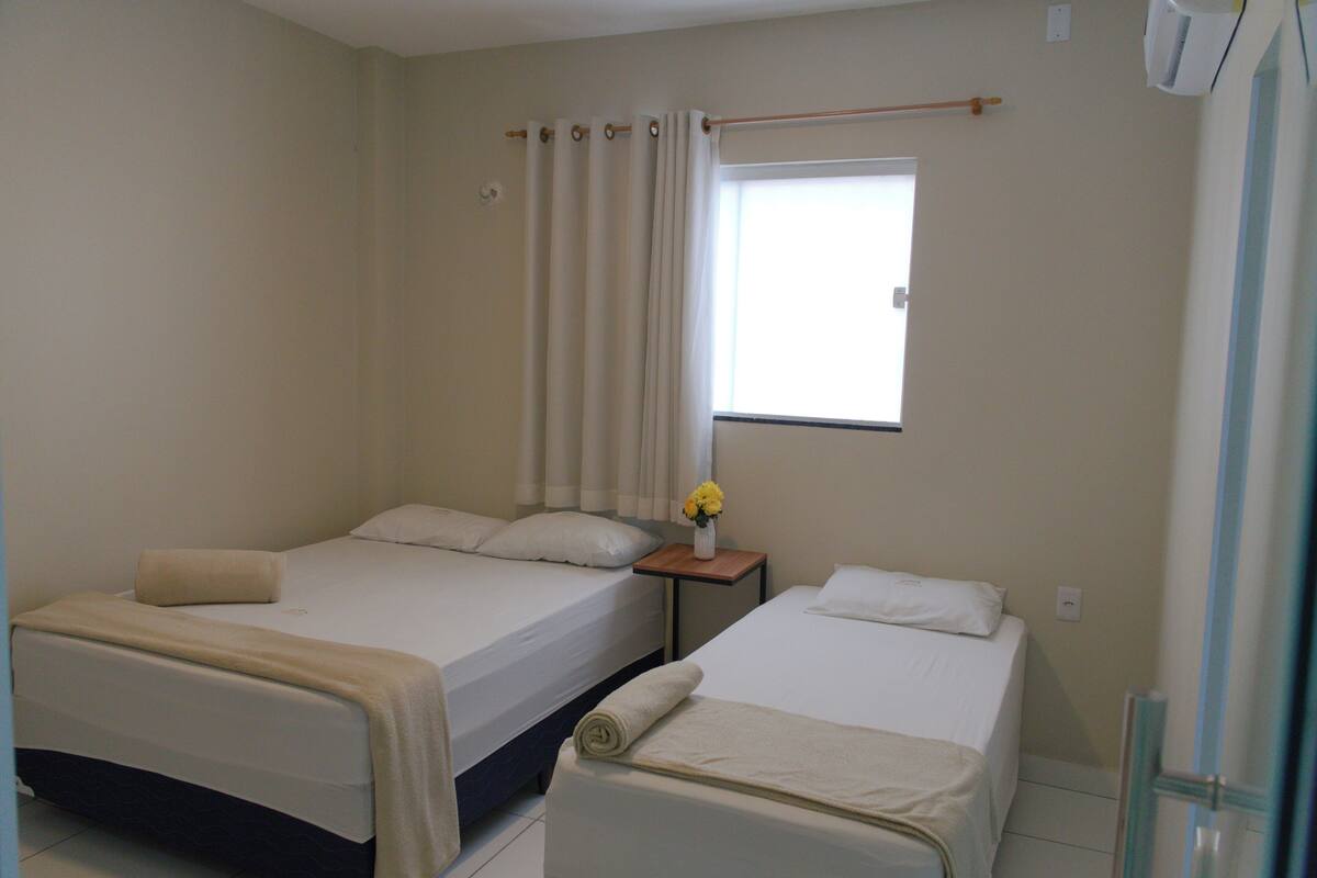 A hotel room with two beds, a window with curtains, a small table with a vase of flowers, and a wall-mounted air conditioner.
