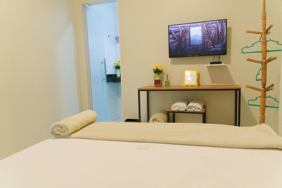 A hotel room with a bed, a television mounted on the wall, a small table with towels and a vase of flowers, and a coat rack.