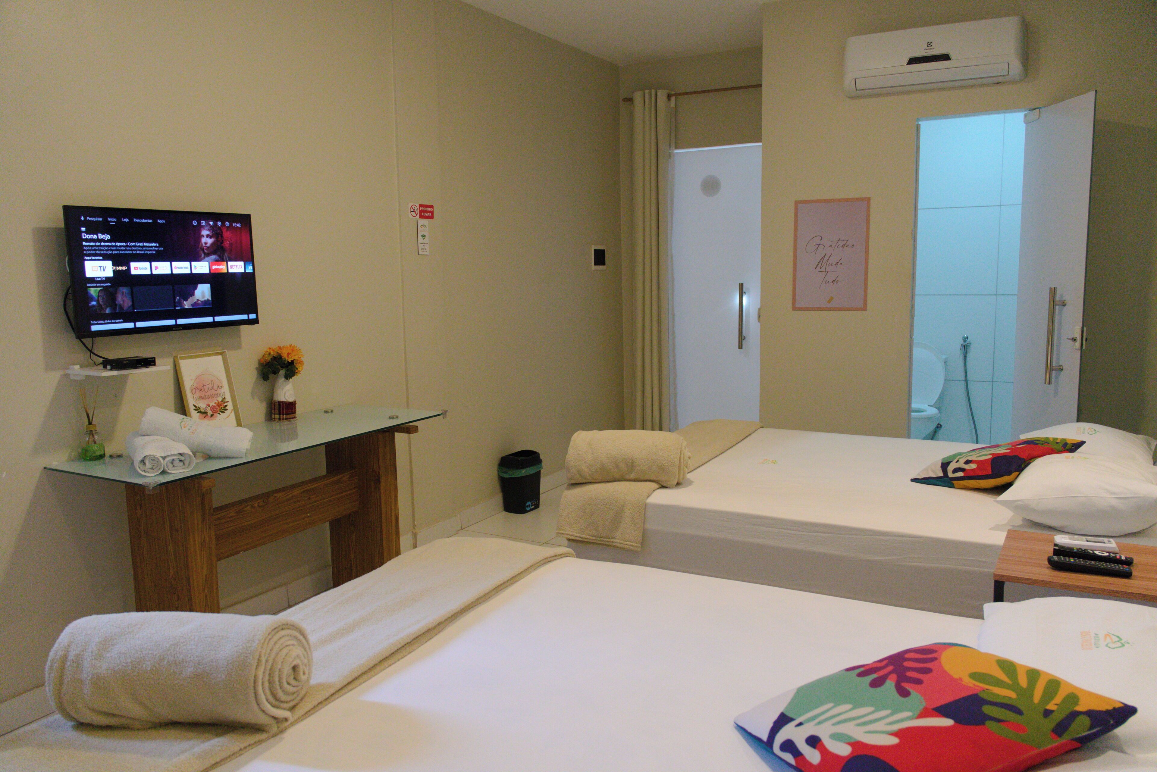 Quarto de hotel com duas camas, televisão fixada na parede, uma secretária pequena com um comando remoto e porta de casa de banho.
