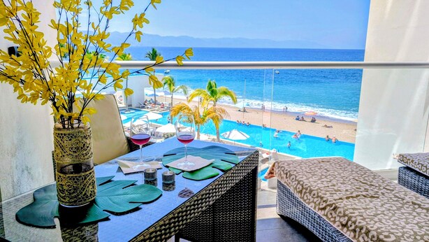 Een balkon met uitzicht op een strand en zwembad, rieten meubilair en een vaas met gele bloemen.