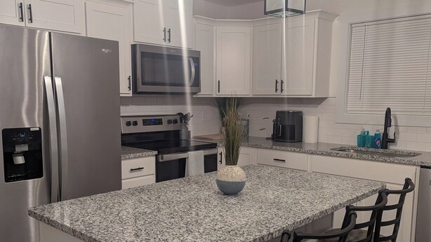 A modern kitchen with white cabinets, a stainless steel refrigerator, a microwave, a stove, and a large granite island with bar stools. - Located in the Eli community of Russell Springs near Beautiful Lake Cumberland. (Russell Springs)