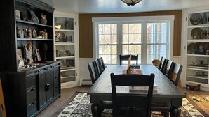 A dining room with a long table, chairs, a patterned rug, and built-in shelving with various items. - Rustic 5-Bedroom Home on 140 Acres perfect for White Mountain family getaways (Campton)
