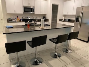 A modern kitchen with white cabinets, a stainless steel refrigerator, a microwave, and a large island with bar stools.
