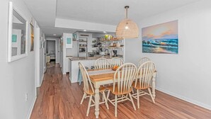 A modern kitchen with white cabinets, a dining area with a wooden table and chairs, and a hanging light fixture. - Direct Oceanfront 3BR/3BA | Upgraded Condo w/ Pool (North Myrtle Beach)