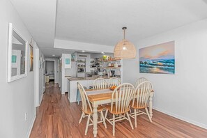 A modern kitchen with white cabinets, a dining area with a wooden table and chairs, and a hanging light fixture. - Direct Oceanfront 3BR/3BA | Upgraded Condo w/ Pool (North Myrtle Beach)