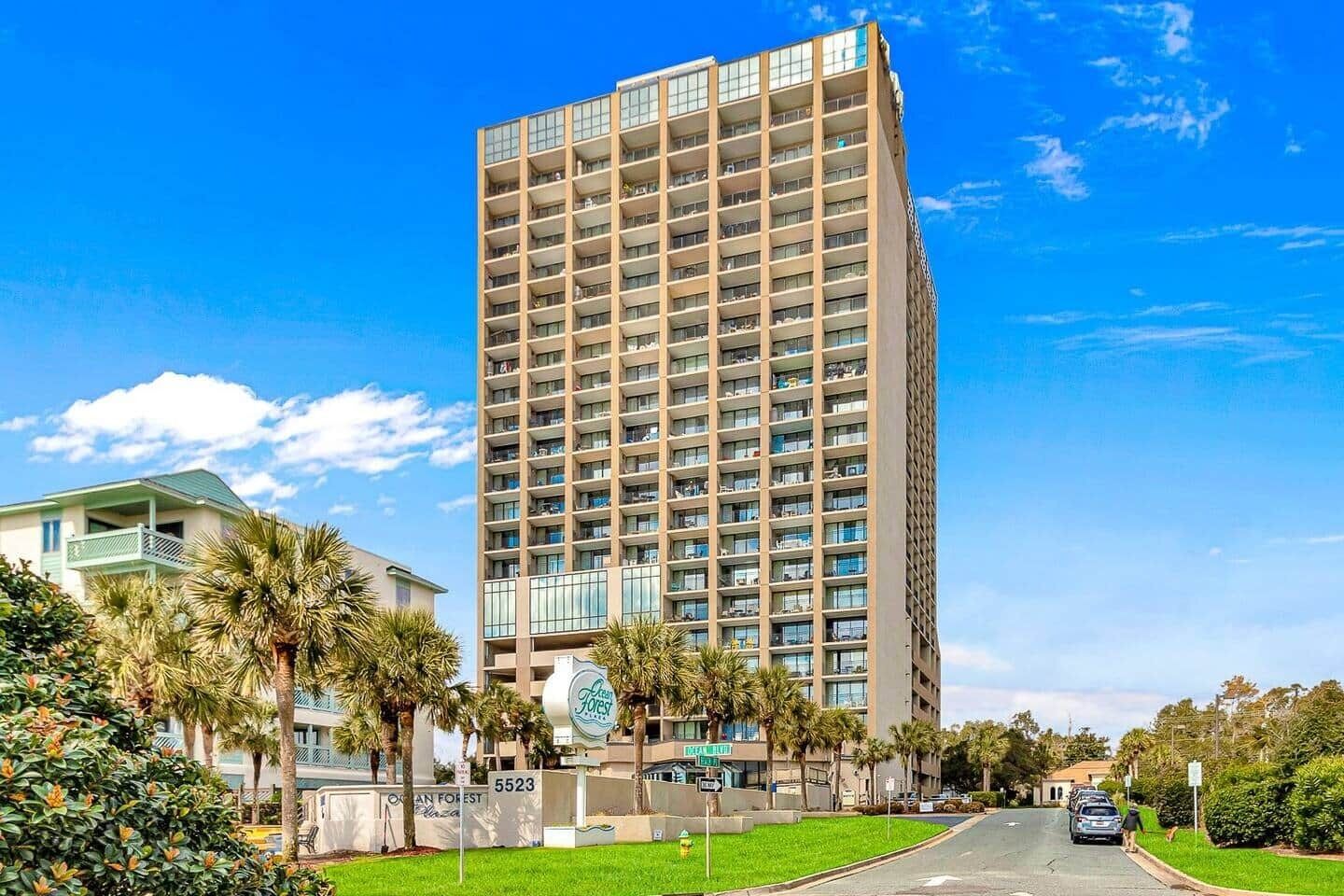 A tall hotel with multiple floors, a sign indicating 'Ocean Forest', and a well-maintained lawn with palm trees.