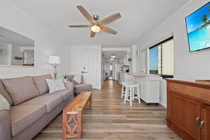 A modern living room with a sofa, a wooden bench, a ceiling fan, and a kitchen area with a window. - Luxury Direct Oceanfront 1BR on the Boardwalk (Myrtle Beach)
