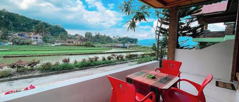 A balcony with a table and chairs, overlooking a view of rice fields and a distant village.