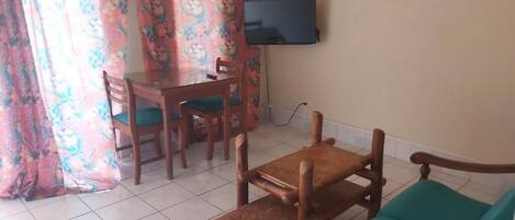 A living room with a sofa, a wooden coffee table, a dining table with chairs, a television mounted on the wall, and a window with curtains.