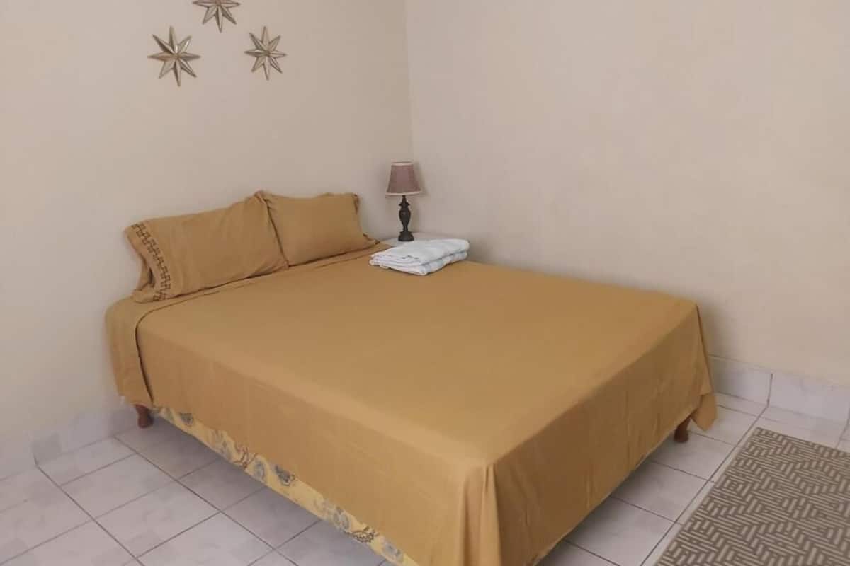 A single bed with a beige bedspread and pillows, a bedside table with a lamp, and a patterned rug on a tiled floor.