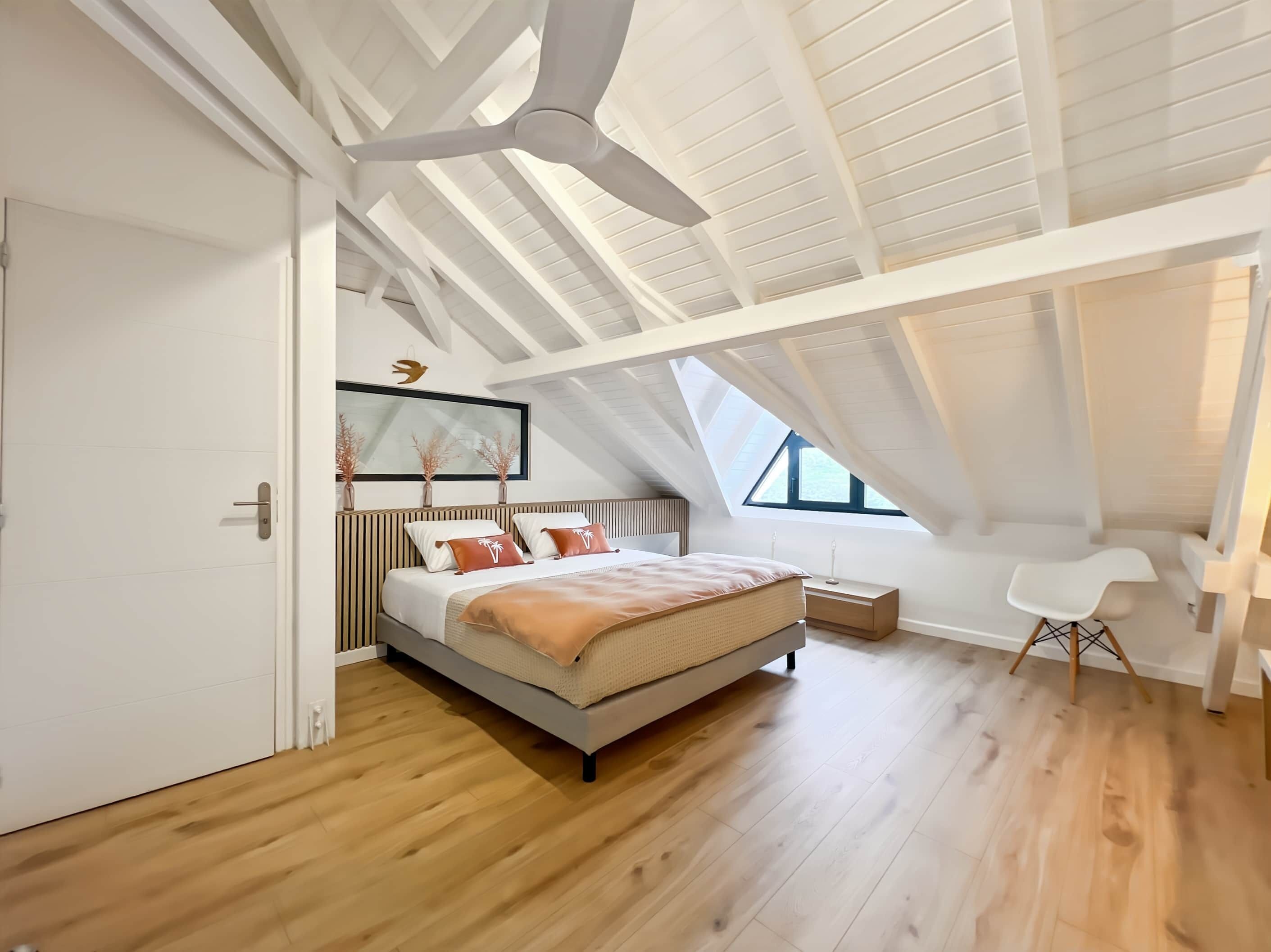 A modern bedroom with a sloped ceiling, a large bed, a bedside table, a chair, and a window.