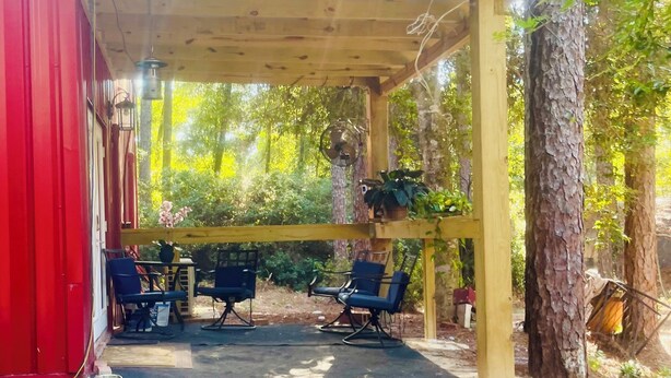 A covered outdoor seating area with chairs and a table, surrounded by trees and a red building.