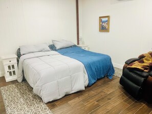 Un dormitorio con cama, mesita de noche, silla, alfombra y un cuadro en la pared.