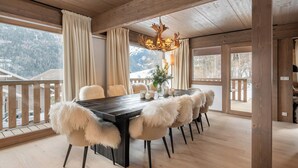 A dining area with a wooden table, chairs with fluffy cushions, a chandelier, and a view of snowy mountains through large windows. - Chalet Quatre Coeurs with sauna in Saint Gervais (Saint-Gervais-les-Bains)