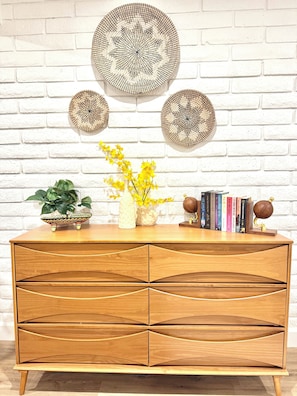 A wooden cabinet with multiple drawers, a vase with yellow flowers, and decorative plates on a white brick wall.