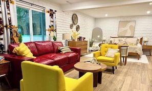 A modern living room with a red leather sofa, yellow armchairs, a wooden coffee table, and a bed with a white headboard.