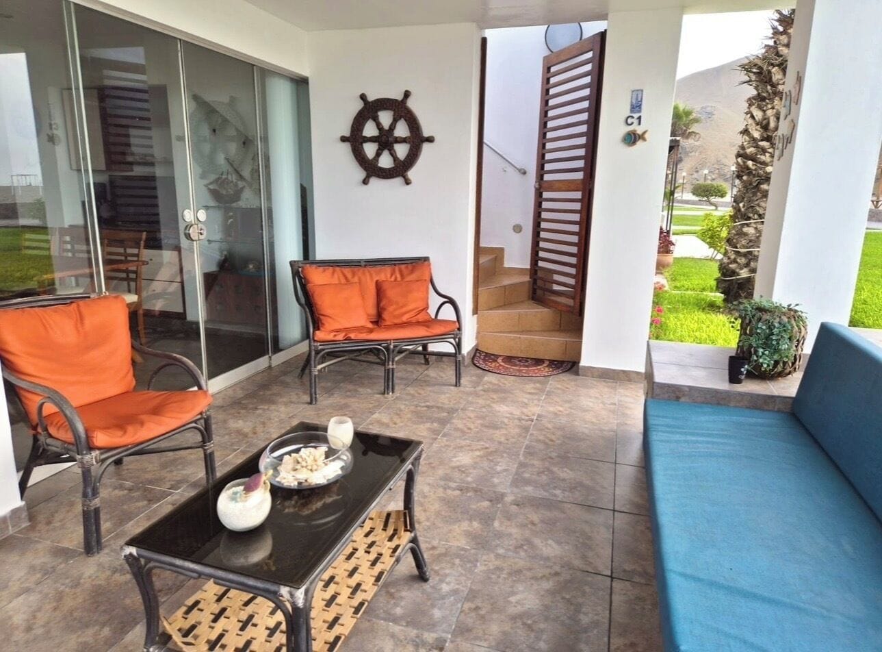 Un patio al aire libre con un sofá azul, dos sillas de mimbre con cojines naranjas, un ventanal de vidrio que da a una habitación y un adorno de timón de barco montado en la pared.