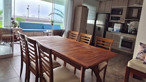 A modern kitchen with wooden cabinets, a dining table with chairs, and a view of the outside through large windows.