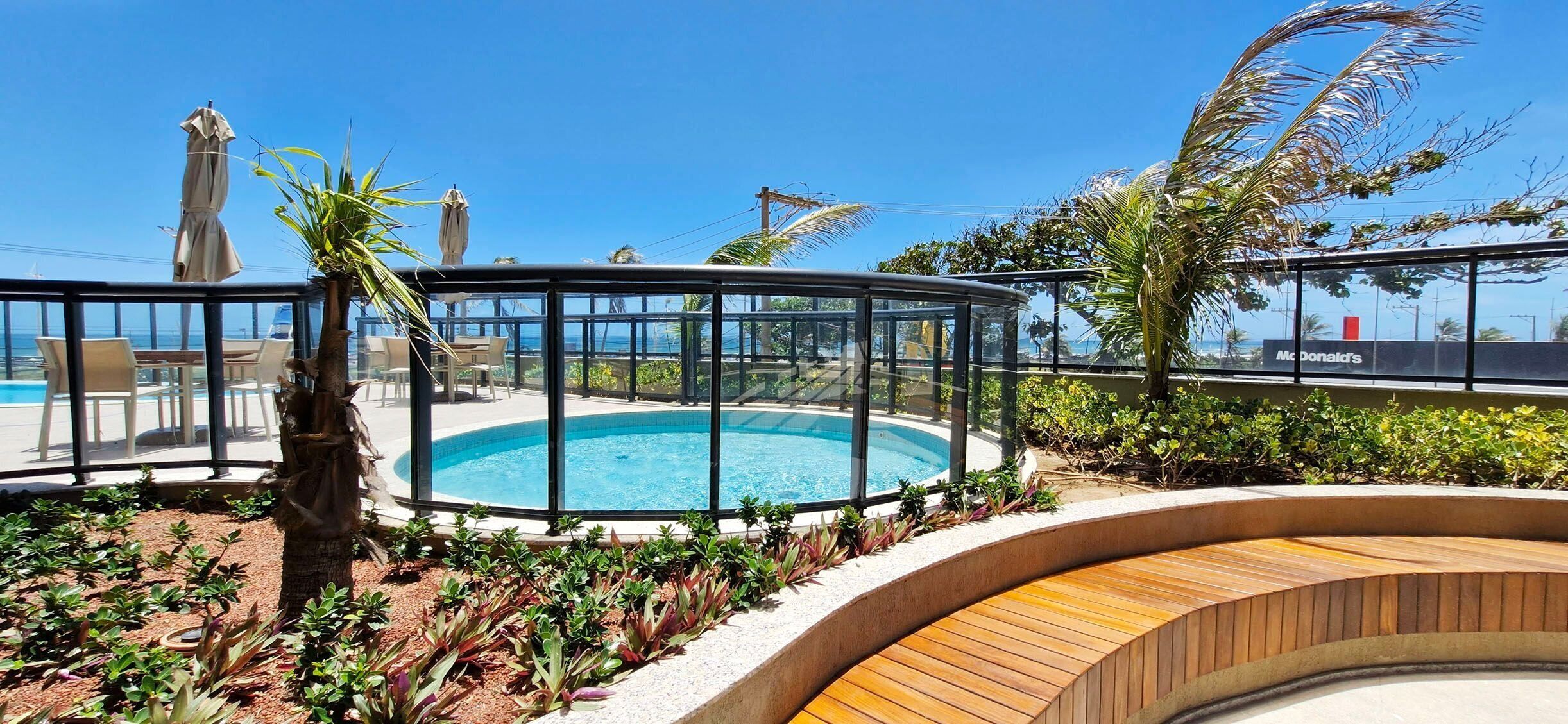 Zona de piscina con cerramiento de vidrio, rodeada de plantas y plataforma de madera.