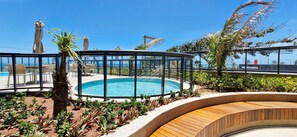 Área de piscina com estrutura de vidro, cercada por plantas e deck de madeira.