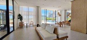 A spacious living area with a white sofa, wooden coffee table, and chairs. Large windows offer views of a swimming pool and buildings. - COJAG0101 - Beach Class Jaguaribe (Salvador)