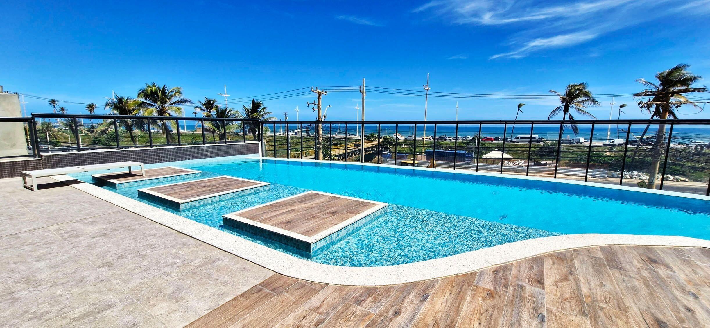 Una piscina con áreas de cubierta de madera, rodeada por una cerca metálica, con vistas a una zona costera con palmeras y un cielo despejado.