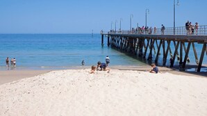 A beach with a pier extending into the water, people walking and sitting on the sand, and a clear blue sky. - Scandi Beach Breeze Minutes from Glenelg Beach (Glenelg)