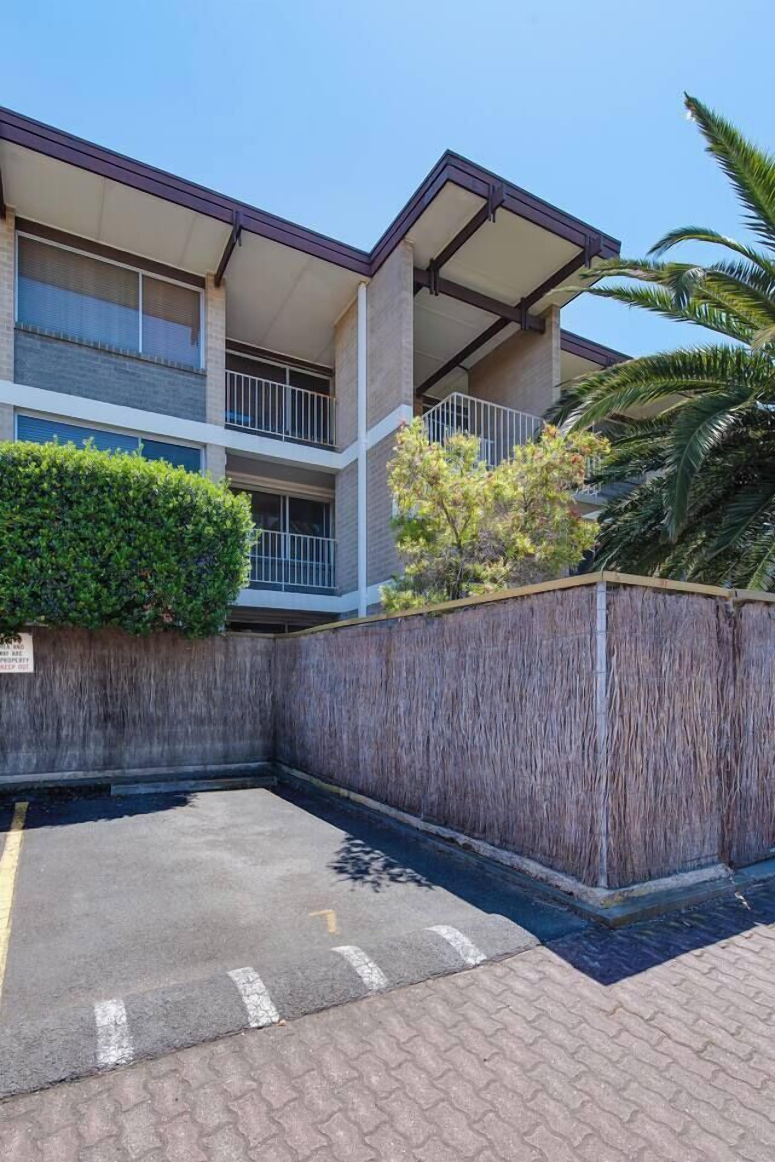 A multi-story building with balconies, a covered parking area, and a wooden fence.