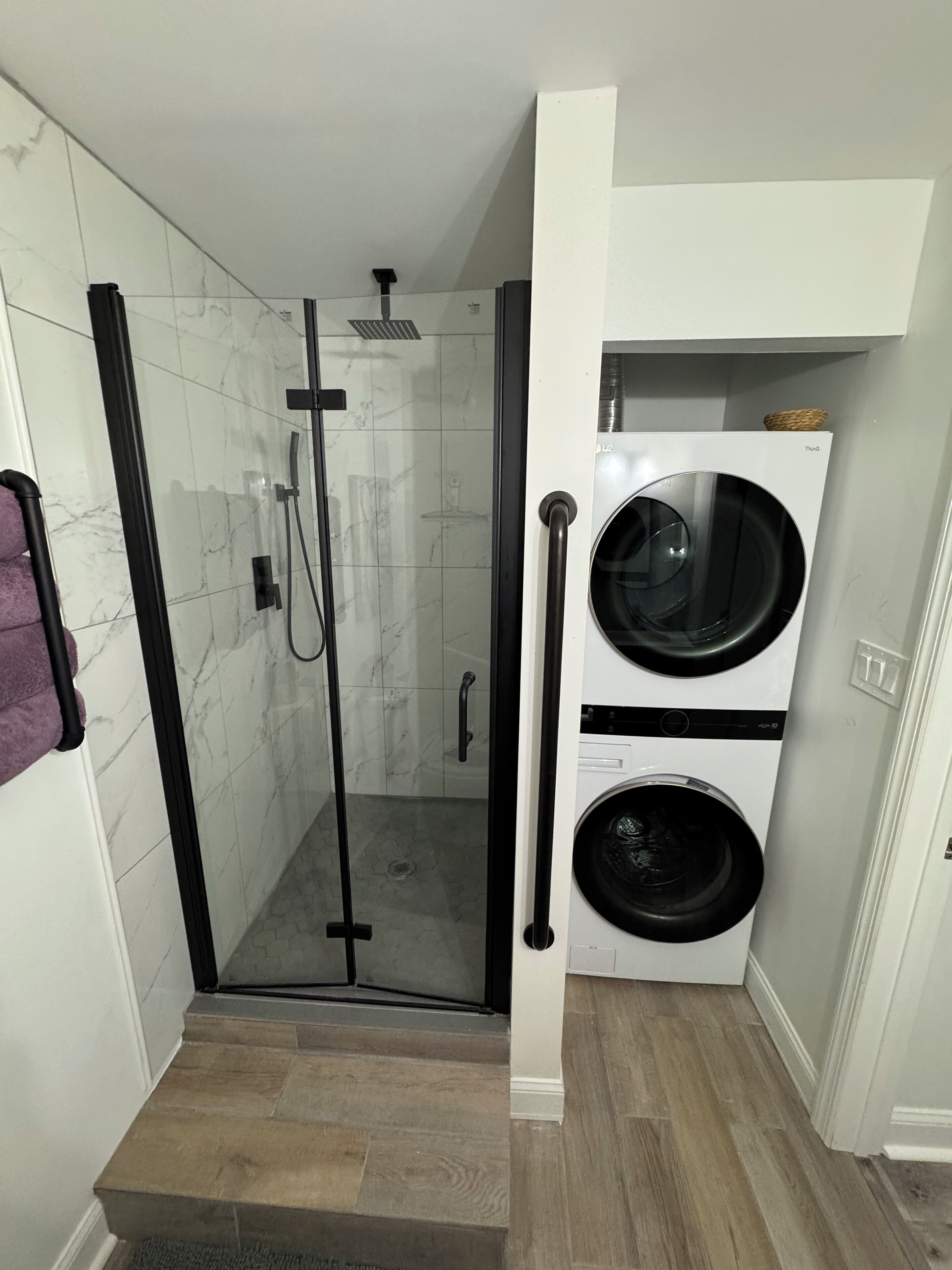 Un baño moderno con ducha envuelta en vidrio, lavadora y secadora integradas, y suelo de madera.