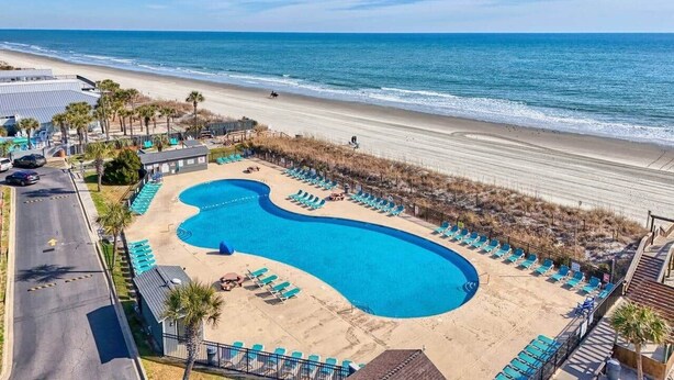 Piscina com design curvo, rodeada por espreguiçadeiras, uma praia e céu claro.
